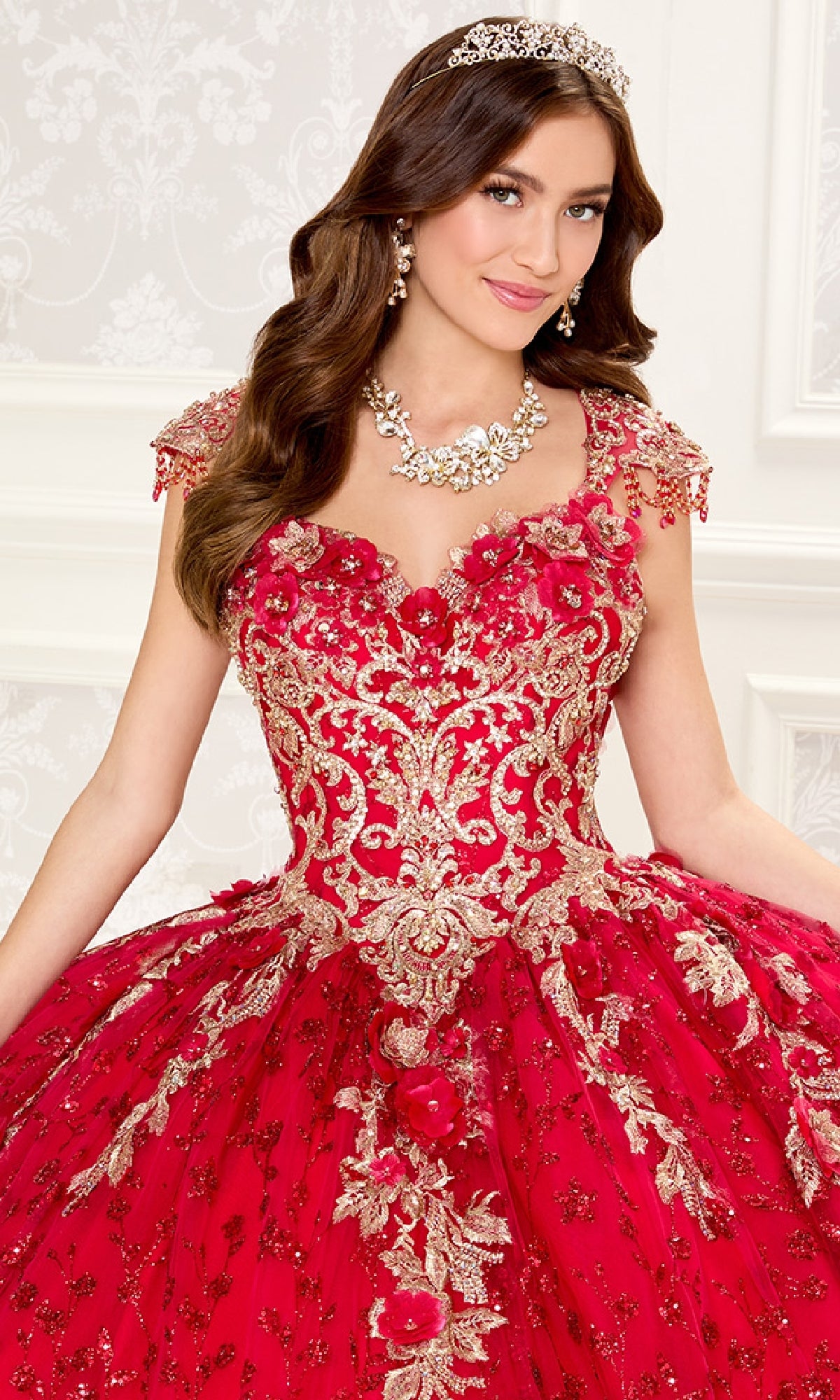A woman in the Quinceanera Dress PR30088 By Princesa, featuring red and gold lace embroidery, stands with a tiara, earrings, and necklace against a decorative white background.